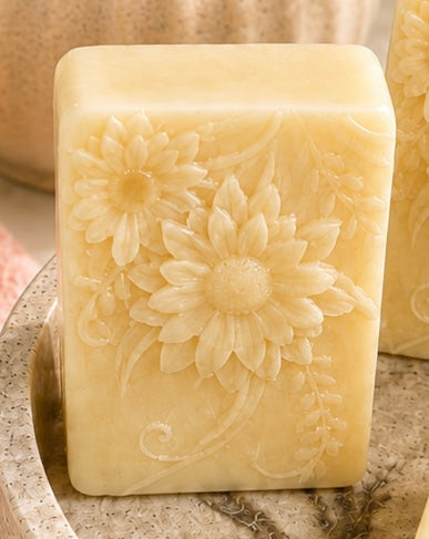 A rectangular bar of pale yellow soap with embossed daisy flowers, resting on a speckled stone surface.